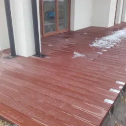 Nowoczesny taras z desek kompozytowych w odcieniach brązu, częściowo pokryty topniejącym śniegiem, z widocznymi elementami konstrukcyjnymi i fragmentem elewacji budynku.