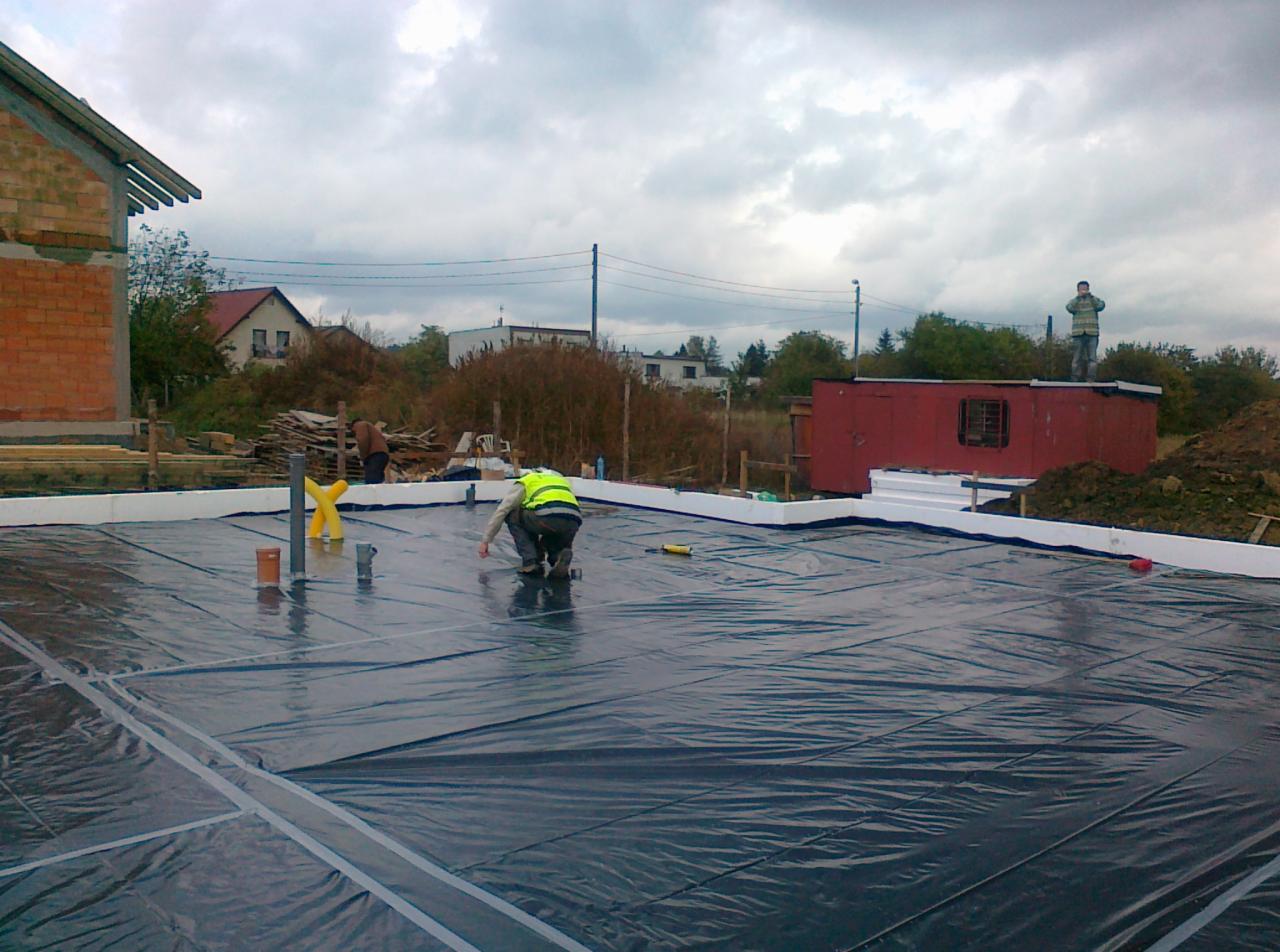 Prace przygotowawcze do wylewki fundamentów: robotnik w kamizelce układa folię hydroizolacyjną na styropianie, widoczne rury i elementy instalacji. W tle budowa domu.