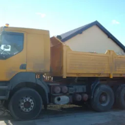 Żółta ciężarówka wywrotka Renault Kerax z pomarańczowym kogutem na dachu kabiny, zaparkowana na betonowym placu, z widocznym fragmentem domu w tle.