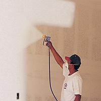 Mężczyzna w czapce i koszulce roboczej natryskuje farbę na ścianę za pomocą pistoletu natryskowego, tworząc równomierną powłokę. Widoczny kabel od pistoletu. Świeżo pomalowana część ściany...