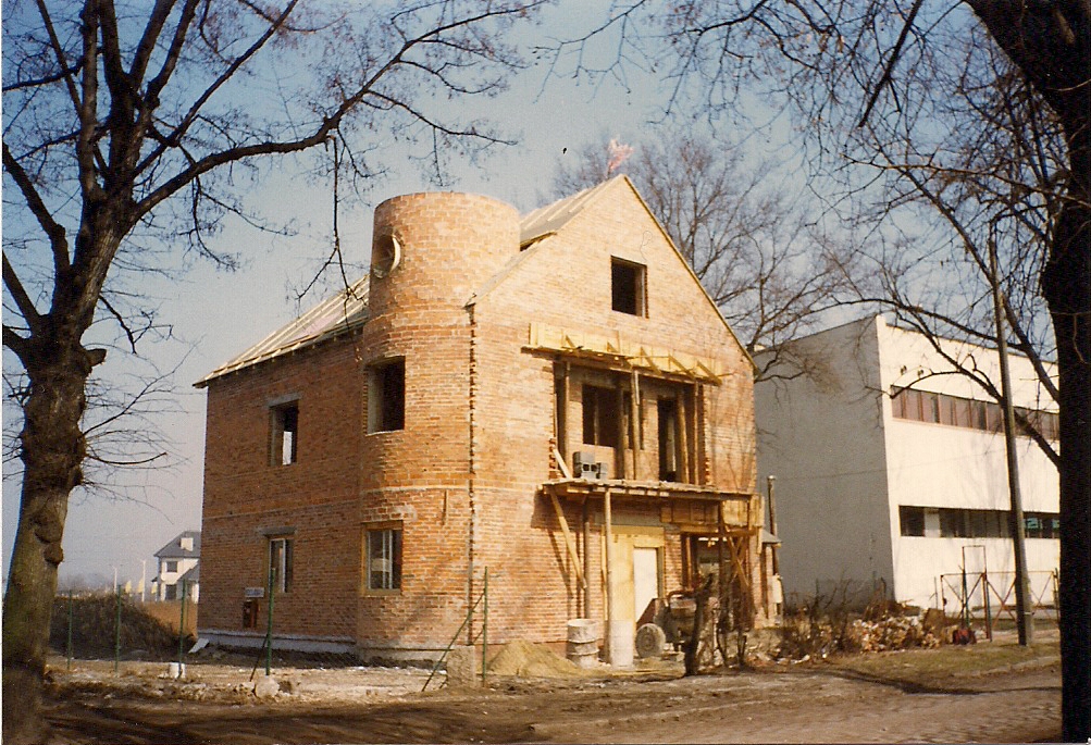 Budynek w trakcie wznoszenia z czerwonej cegły, z charakterystyczną okrągłą wieżyczką, widoczne drewniane elementy konstrukcyjne i rusztowania, na pierwszym planie drzewa bez liści, w tle inny...