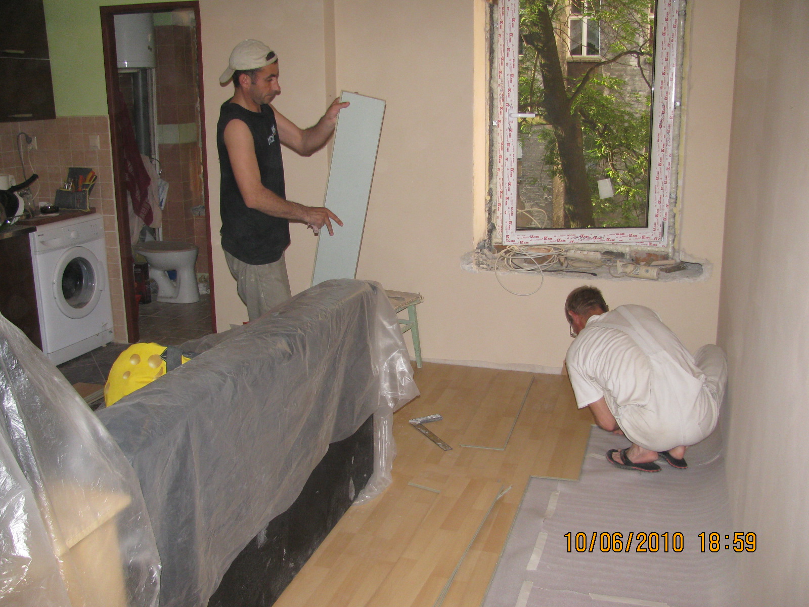 Dwóch mężczyzn podczas układania paneli podłogowych w pokoju z oknem, widoczny fragment kuchni z pralką, meble zabezpieczone folią.