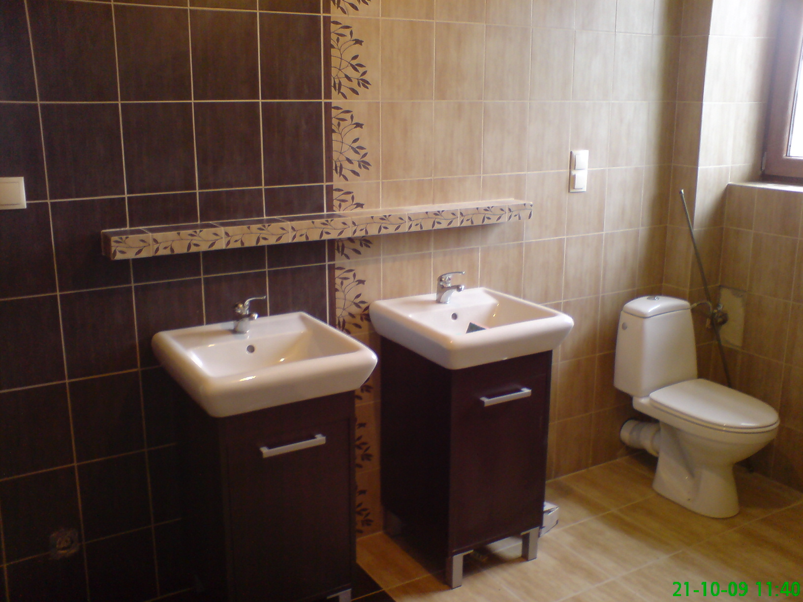 Ukończona łazienka z dwoma umywalkami na ciemnobrązowych szafkach, toaleta z widoczną rurą doprowadzającą wodę, brązowe płytki z dekoracyjnym motywem roślinnym i półką z tym samym motywem.