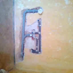 Fragment ściany z odsłoniętą instalacją hydrauliczną z szarych rur, widoczne połączenia i zawory, ściana w kolorze żółtym z odpryskami farby, w tle fragment podłogi.