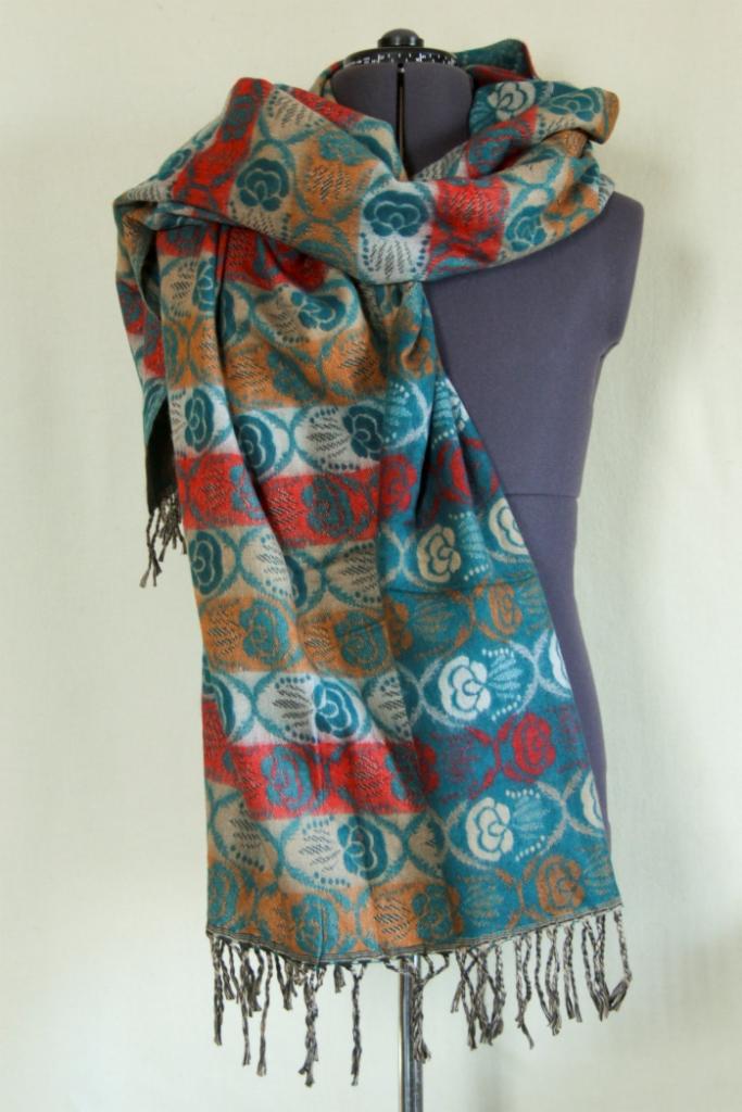 Wzorzysty szal z frędzlami w odcieniach turkusu, czerwieni, beżu i pomarańczy, ułożony na szarym manekinie krawieckim.