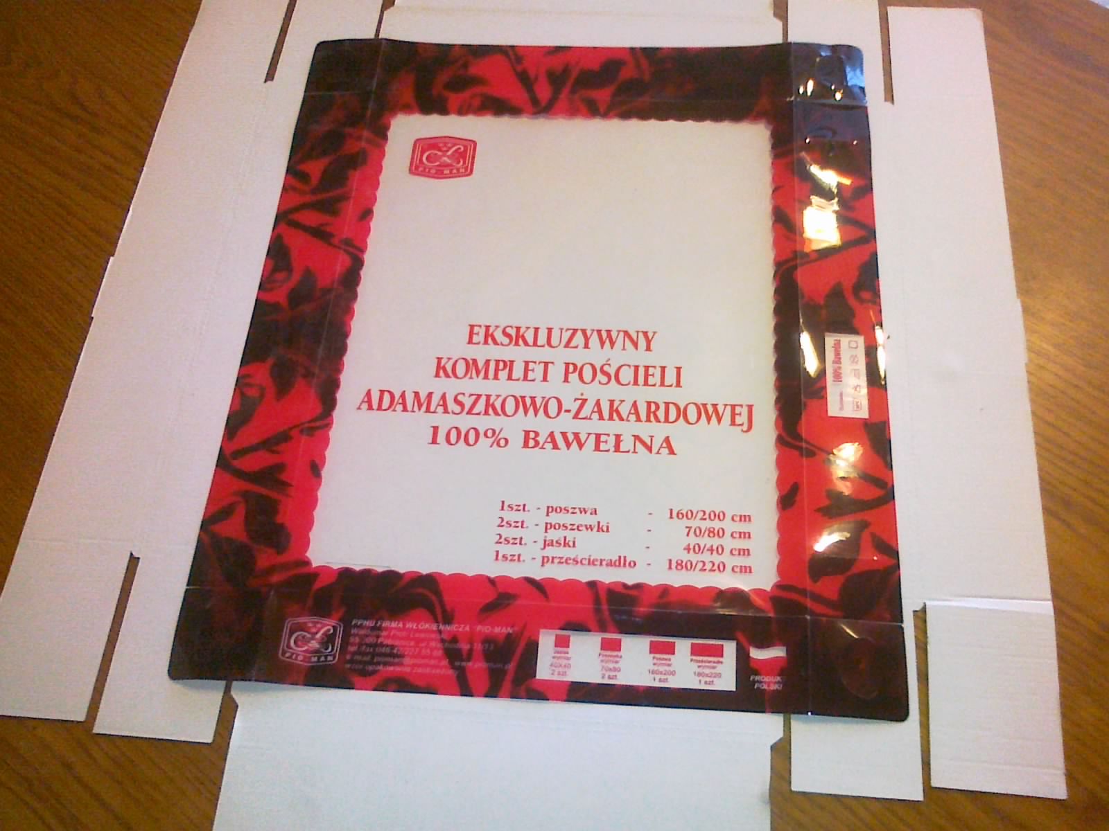 Rozłożone kartonowe opakowanie z nadrukowanym wzorem i informacjami o ekskluzywnym komplecie pościeli adamaszkowo-żakardowej ze 100% bawełny, leżące na drewnianym stole.