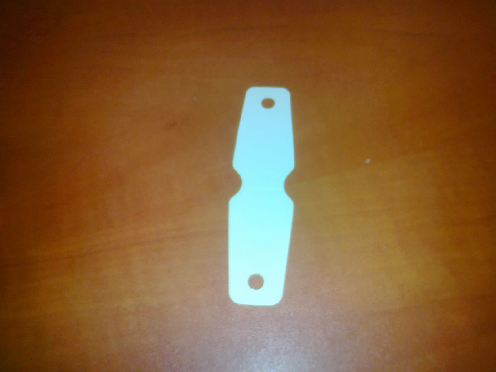 Biała, kartonowa zawieszka do biżuterii z dwoma otworami, leżąca na brązowym, drewnianym blacie.