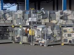 Metalowe kosze wypełnione zużytym sprzętem elektronicznym, widoczne monitory, komputery i inne urządzenia, ustawione na zewnątrz budynku.