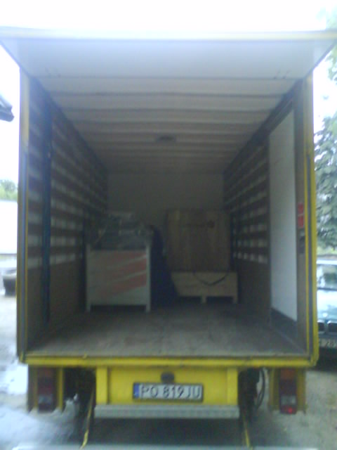 Wnętrze żółtego samochodu dostawczego z otwartymi drzwiami, widoczne zabezpieczone kartonami przedmioty gotowe do transportu, tablica rejestracyjna PO 812JU.