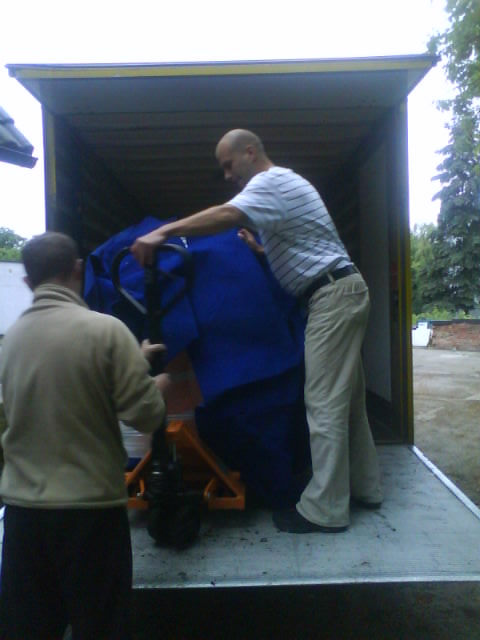 Dwóch mężczyzn ładuje mebel owinięty w niebieski materiał do wnętrza furgonetki za pomocą wózka paletowego.