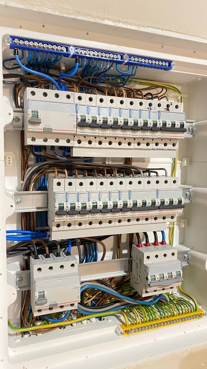 Rozdzielnica elektryczna Legrand z precyzyjnie ułożonymi przewodami w kolorach: niebieskim, brązowym i żółto-zielonym. Widoczne zabezpieczenia nadprądowe i szyna wyrównawcza.