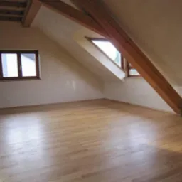 Jasne panele podłogowe w pokoju na poddaszu z drewnianymi belkami i oknami. Ściany w odcieniach beżu. Widok na gotowe wnętrze po remoncie.