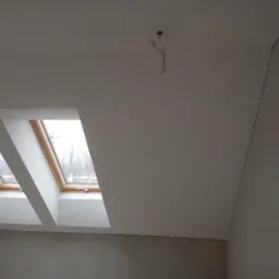 Poddasze po remoncie: Dwa okna dachowe z drewnianą ramą, świeżo pomalowany sufit i ściany w odcieniach bieli, widoczne kable wystające z sufitu, listwy przypodłogowe zamontowane.