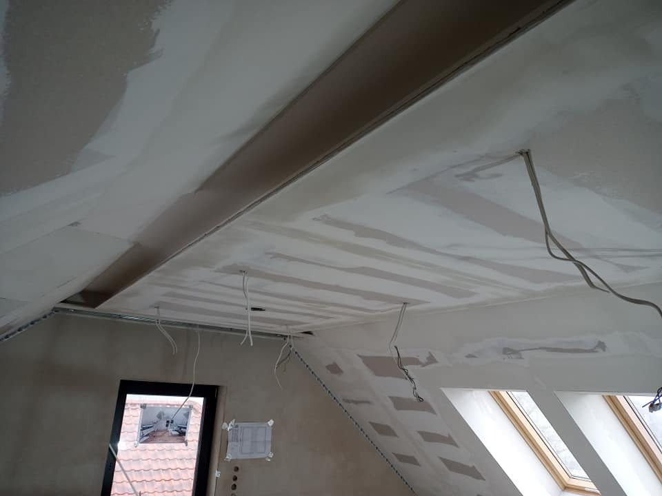 Poddasze w trakcie prac wykończeniowych: widoczne płyty na suficie, częściowo pokryte masą szpachlową, wystające przewody elektryczne, okno dachowe i fragment ściany ze śladami wiercenia.