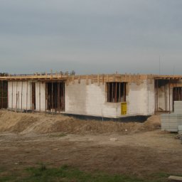 Roman Puzio - Wznoszenie ścian domu z bloczków betonu komórkowego; widoczne zbrojenia pionowe, drewniany szalunek stropu oraz sterta bloczków na paletach obok budynku.