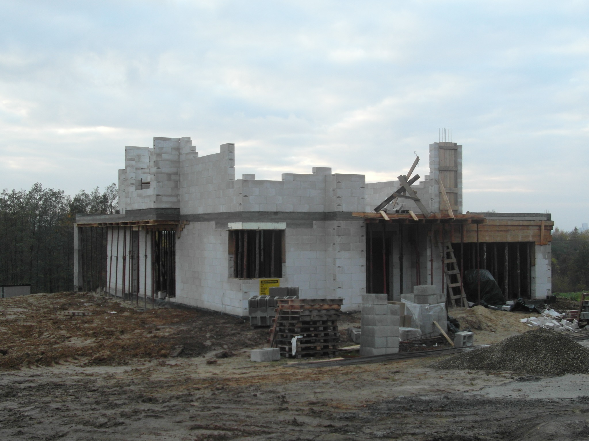Surowy stan domu w trakcie budowy z betonu komórkowego; widoczne ściany, drewniane szalunki, rusztowania i materiały budowlane na błotnistym terenie.
