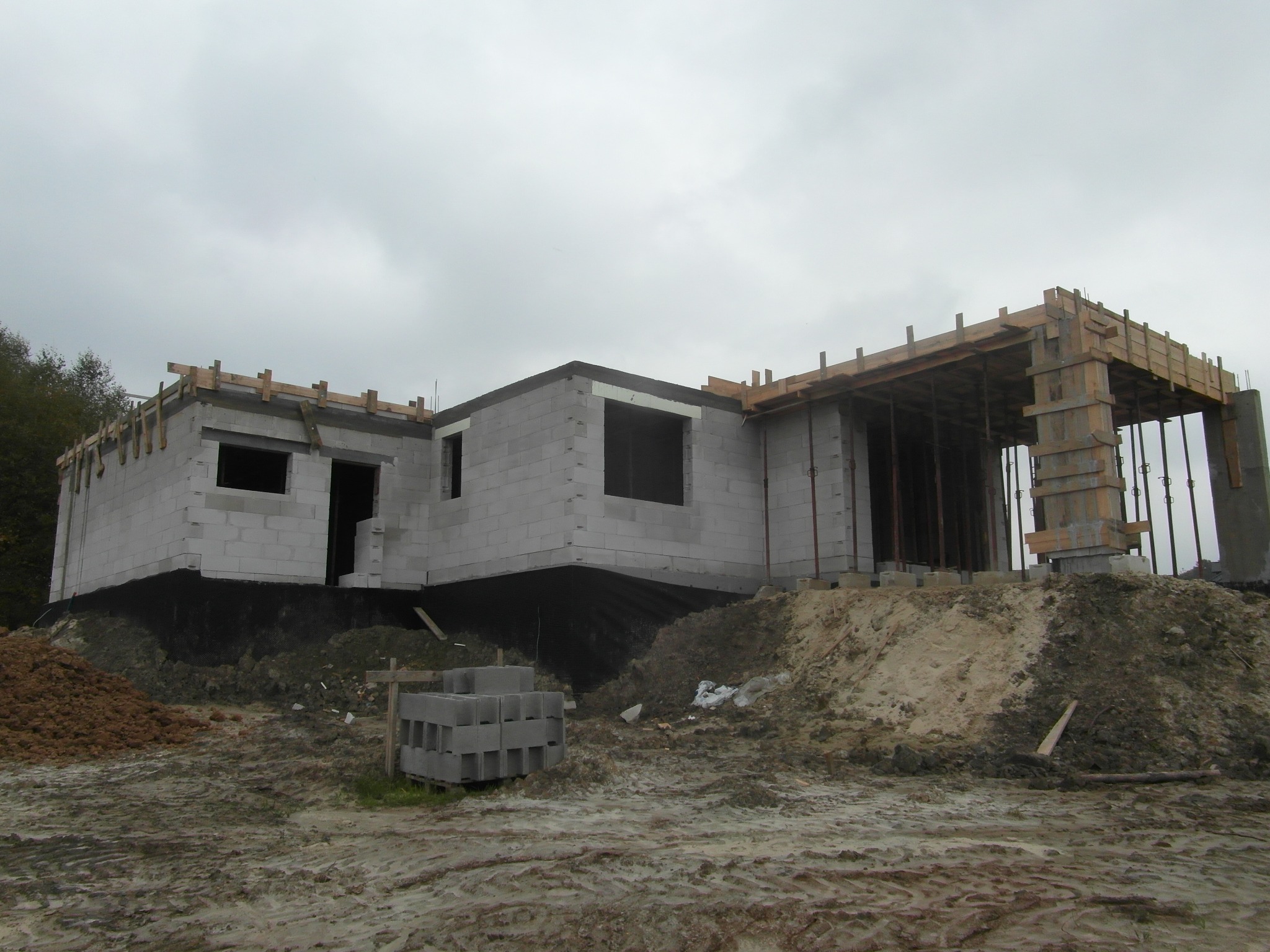 Surowy stan domu z bloczków betonowych w trakcie budowy, widoczne szalunki na stropie i fundamenty zabezpieczone folią, teren wokół placu budowy pokryty błotem i piaskiem.