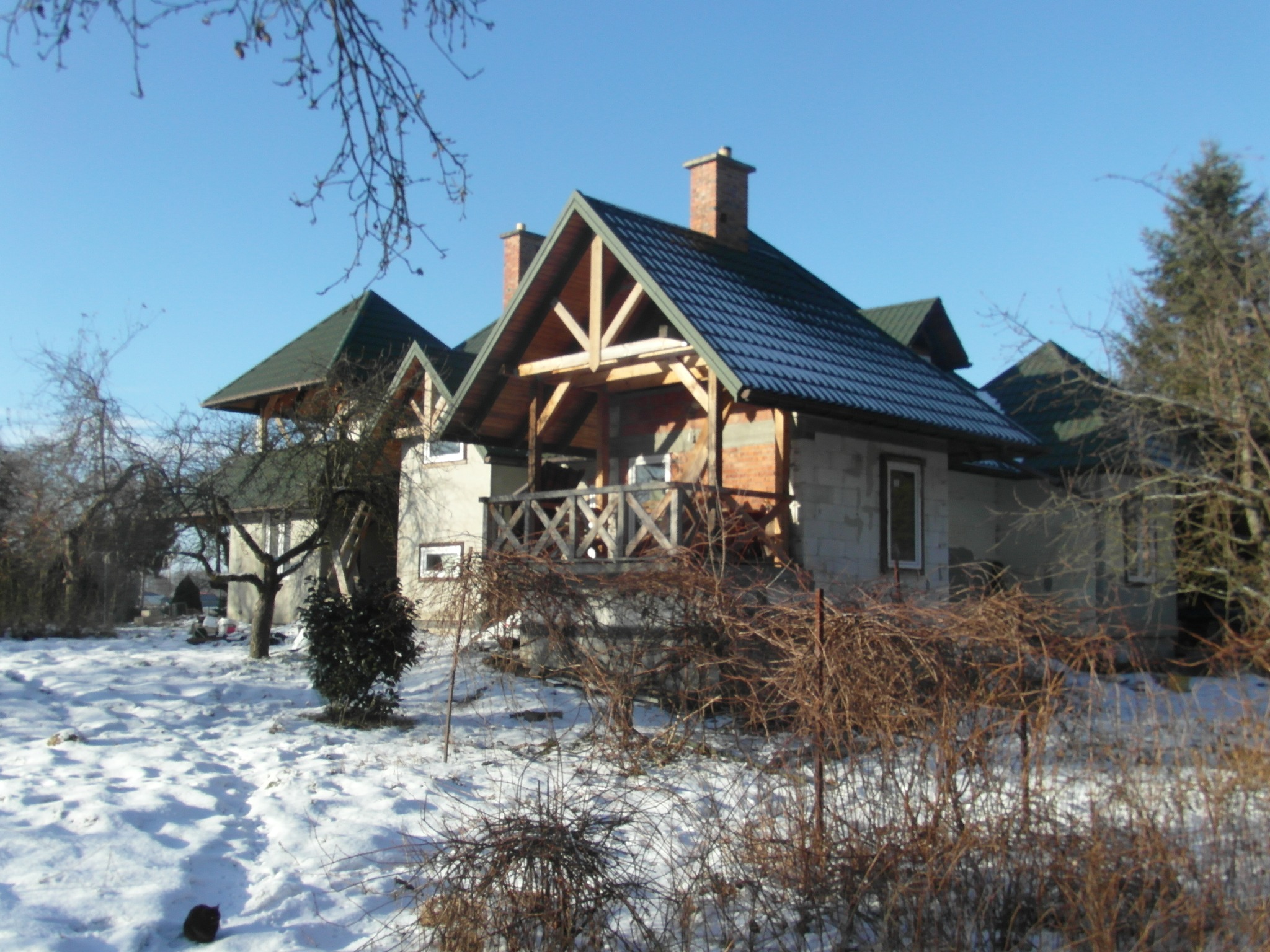 Dom w trakcie budowy, widoczne ściany z bloczków, drewniana konstrukcja dachu i kominy. Zima, śnieg na ziemi, błękitne niebo w tle.