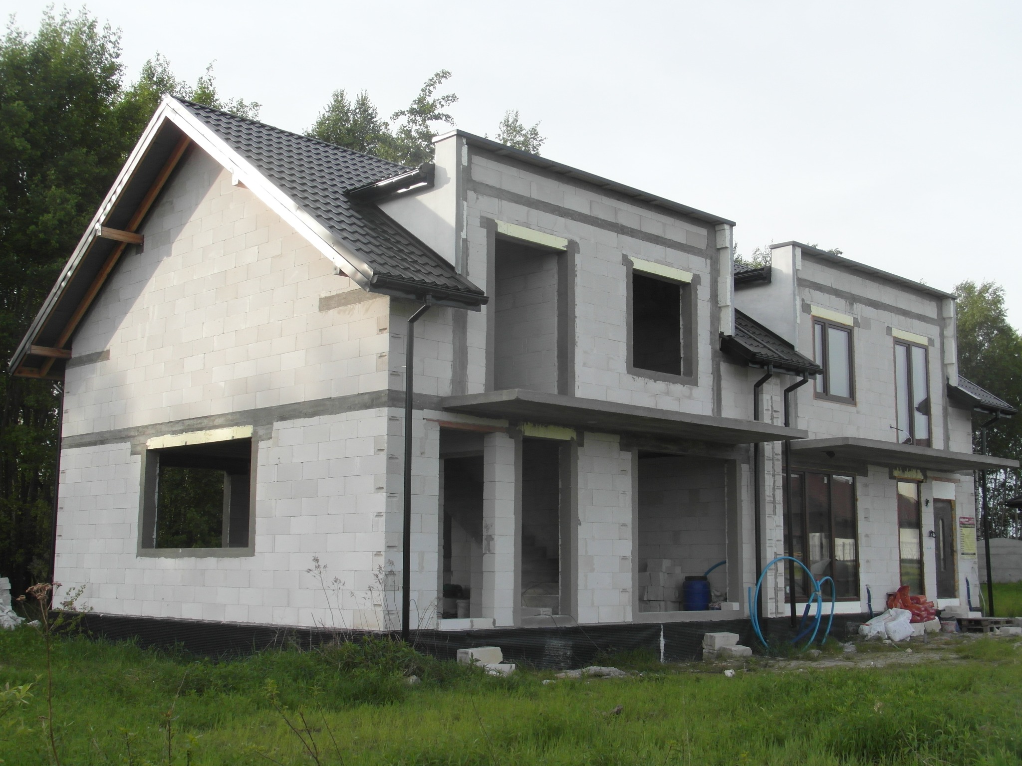 Niedokończony, dwukondygnacyjny dom z białego bloczka, z czarnym dachem. Widoczne otwory okienne i drzwiowe, częściowo zadaszony taras. W tle zieleń drzew.