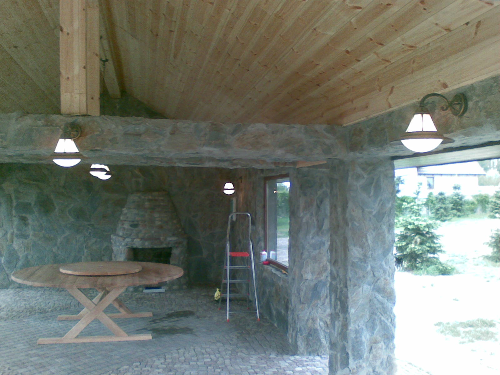 Zadaszona altana z kamienia i drewna z oświetleniem, drewnianym stołem i kominkiem z kamienia, widok na zewnątrz przez okno.