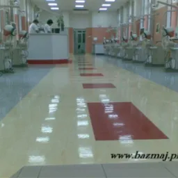 Błyszcząca, świeżo umyta podłoga w dużej sali z wieloma stanowiskami dentystycznymi, widoczne odbicia światła na powierzchni, w tle osoby przy recepcji.