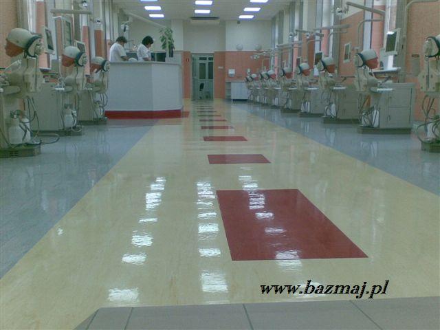 Błyszcząca, świeżo umyta podłoga w dużej sali z wieloma stanowiskami dentystycznymi, widoczne odbicia światła na powierzchni, w tle osoby przy recepcji.