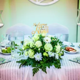 Udekorowany stół weselny z białymi kwiatami, świecami w szklanych świecznikach na wysokich nóżkach i złotym napisem 'Najlepsze dopiero przed Nami'.