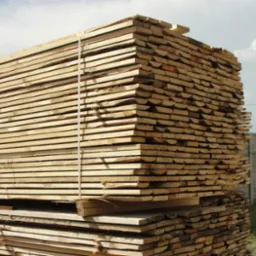 Stosy jasnego drewna budowlanego, zabezpieczone białymi taśmami, ułożone na paletach na tle betonowego ogrodzenia i pochmurnego nieba.