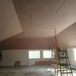 Montaż ocieplenia poddasza z widocznymi płytami na suficie, drabiny, okna z luksferami i pracownik w tle.