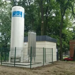 Zabezpieczony zbiornik gazu Linde na terenie zielonym, otoczony ogrodzeniem. Widoczne elementy instalacji i budynek. Dwa samochody zaparkowane w pobliżu.