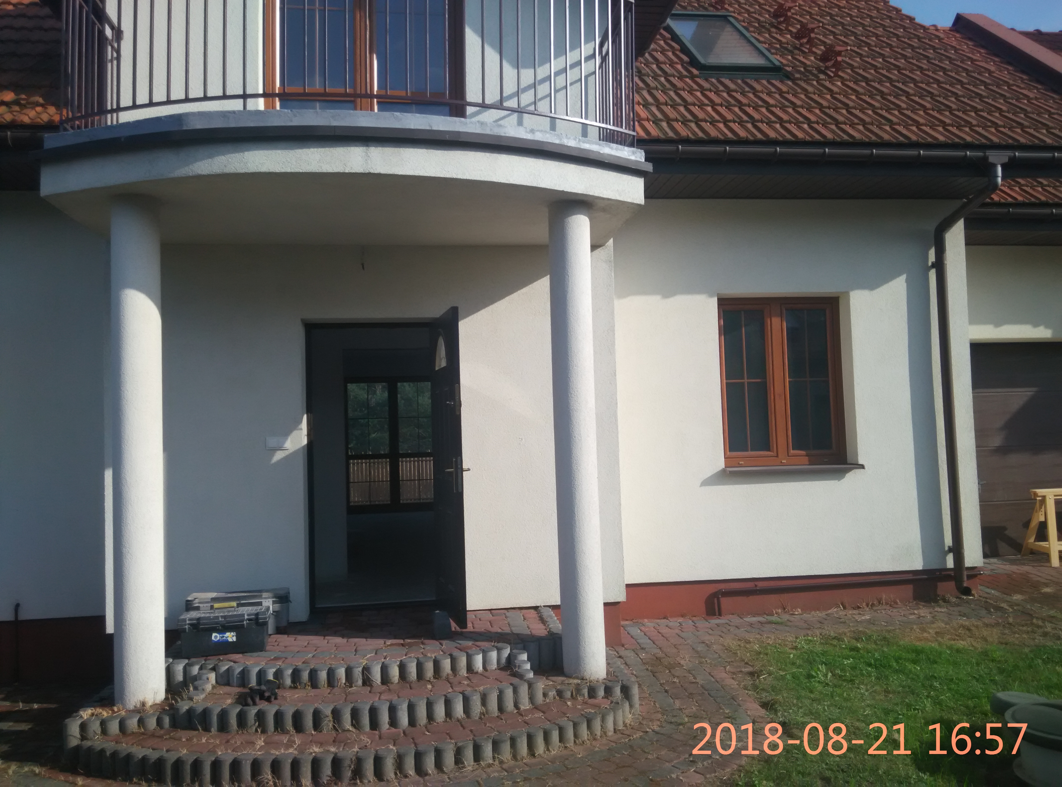 Wejście do domu z kolumnami i schodami z kostki, skrzynka narzędziowa obok schodów, okno z brązową ramą na ścianie.