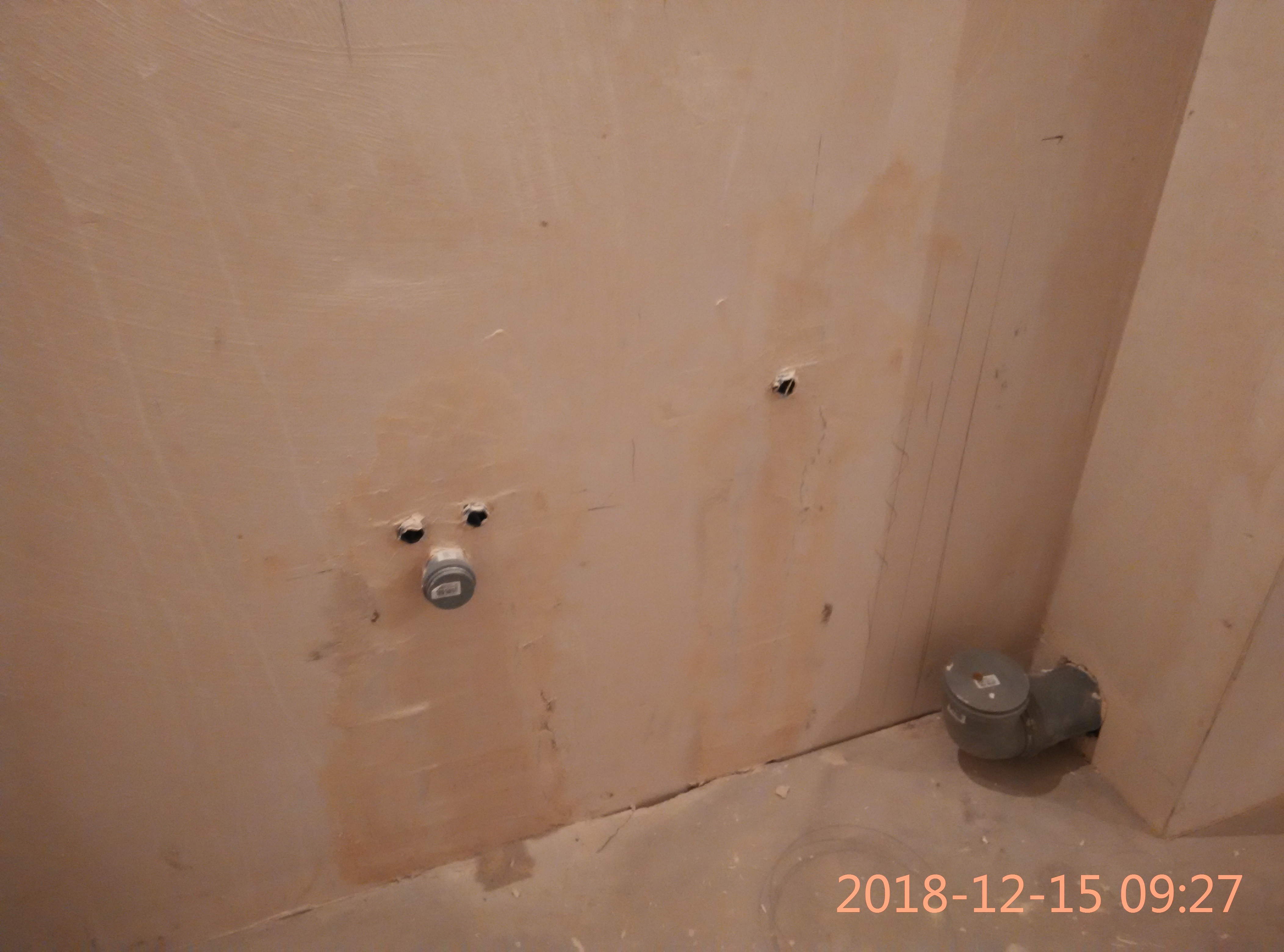 Surowa ściana w trakcie prac hydraulicznych, widoczne dwa otwory w ścianie z wystającymi rurami, jeden otwór z szarą zaślepką, oraz szara rura odpływowa pod kątem na podłodze w rogu pomieszczenia.