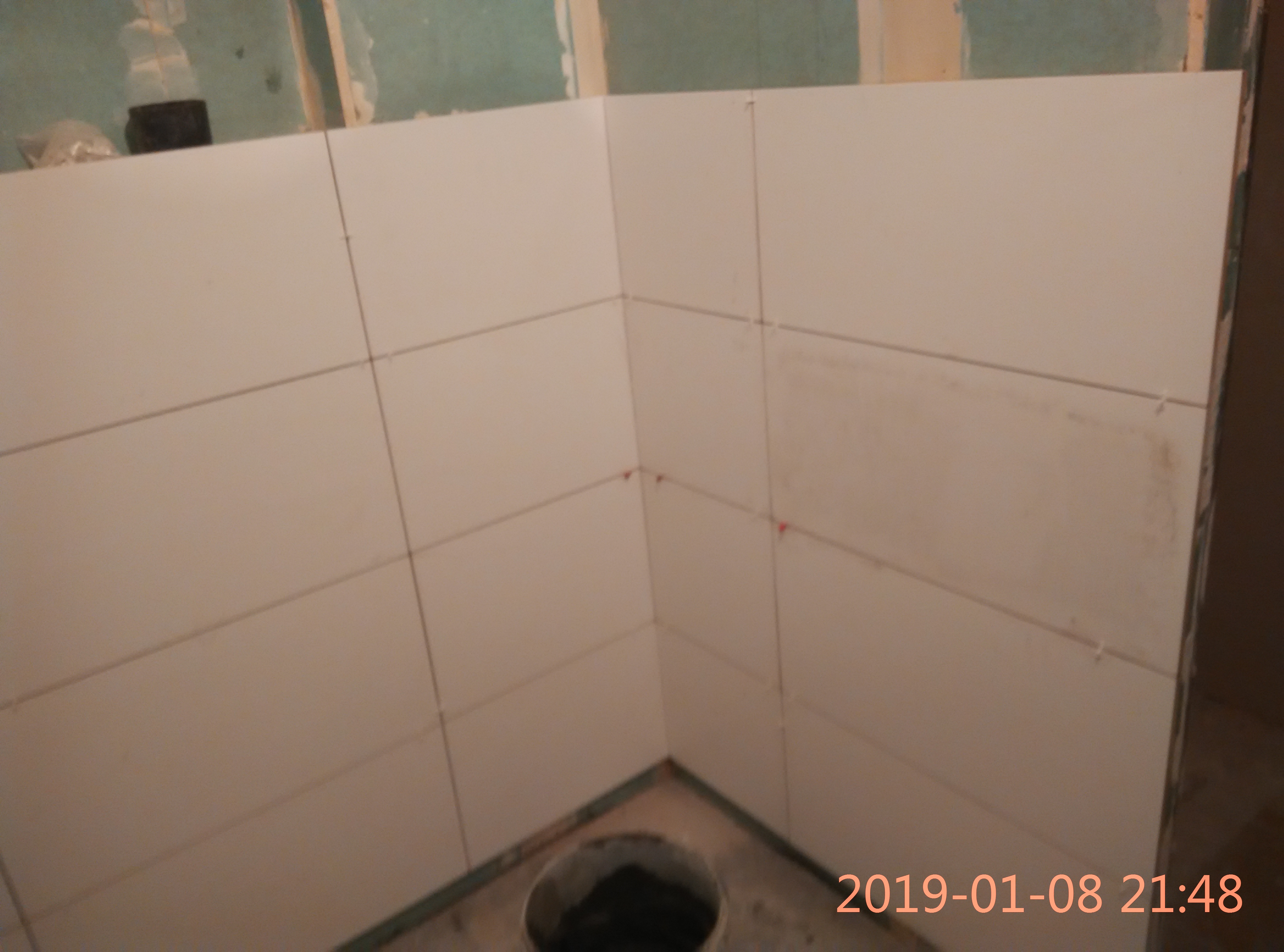 Świeżo ułożone, białe płytki ścienne w rogu łazienki, z widocznymi krzyżykami dystansowymi, niedokładnościami oraz śladami zaprawy, z wiadrem na dole kadru i ścianami w tle przygotowanymi...