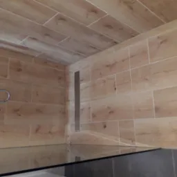 Wykończona łazienka z drewnopodobnymi płytkami na ścianach i suficie, widoczny odpływ liniowy i fragment szklanej kabiny prysznicowej.