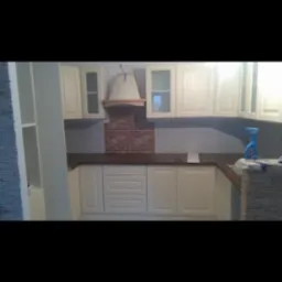 Kremowa zabudowa kuchenna z drewnianym blatem i okapem nad ceglanym panelem, widoczna butelka z płynem czyszczącym na blacie.