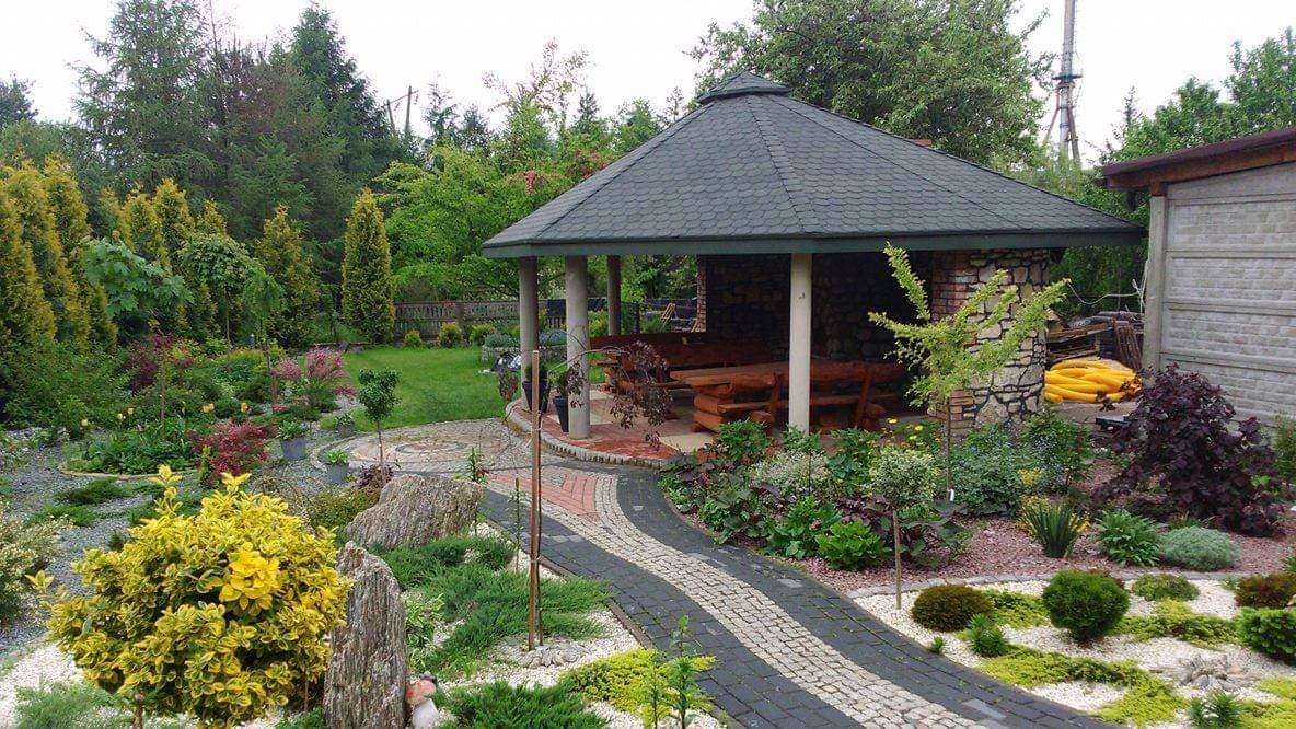 Ogród z altaną z kamienia i drewna, ścieżka z kostki brukowej w dwóch kolorach, rabaty z różnorodną roślinnością, trawnik w tle.