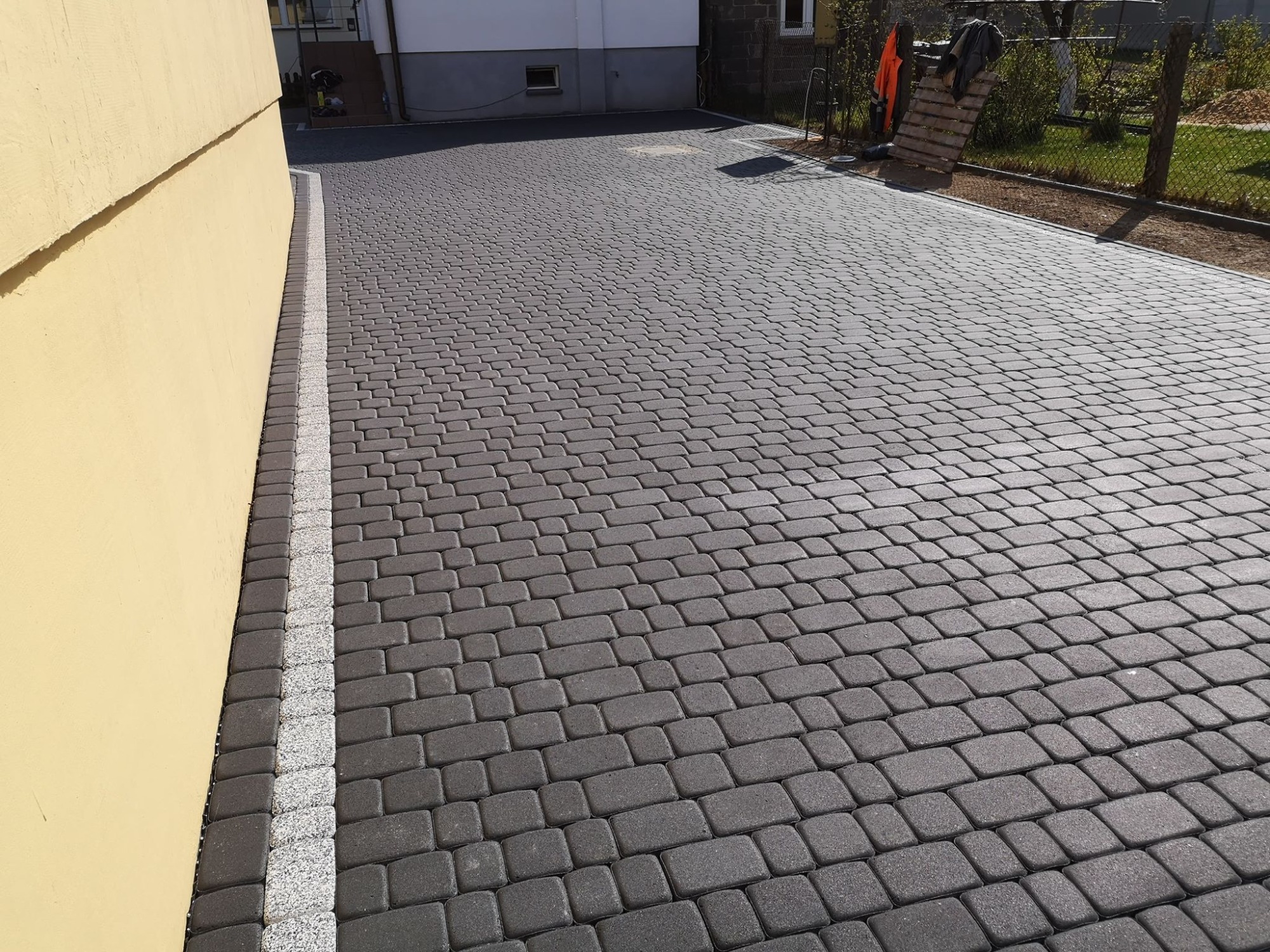 Ułożona kostka brukowa w odcieniach szarości tworząca podjazd, obrzeża z jasnej kostki, widoczny fragment elewacji budynku w tle oraz paleta oparta o ogrodzenie.