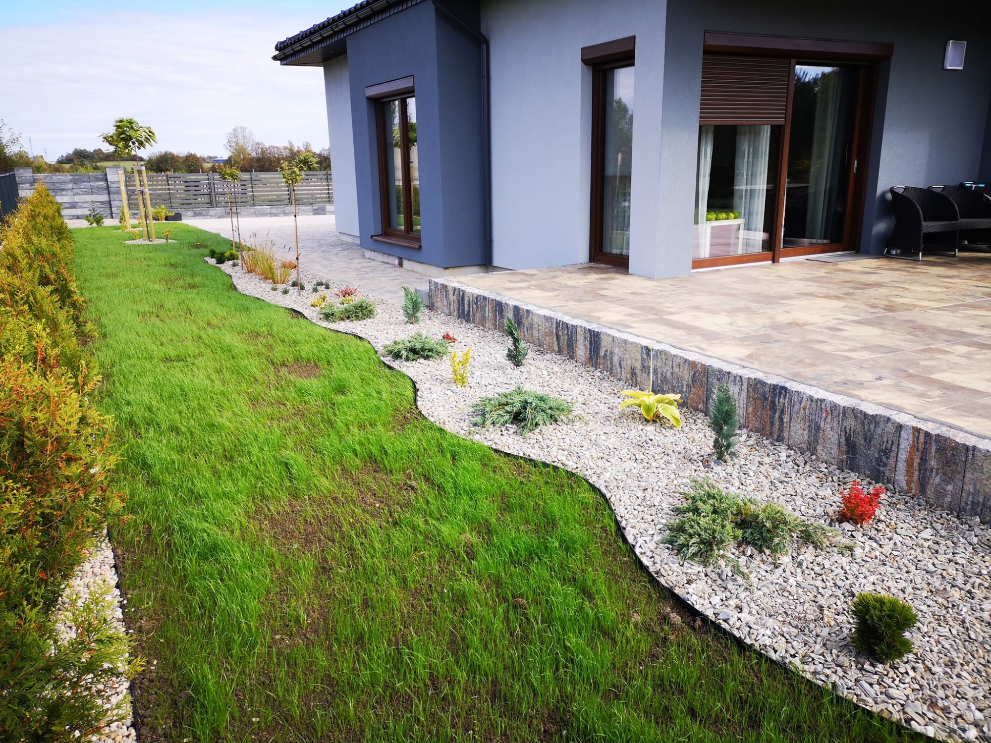 Nowoczesny ogród z trawnikiem, ścieżką z białego żwiru, niskimi roślinami ozdobnymi i kamiennym murkiem oddzielającym ogród od tarasu przylegającego do domu z szarymi elewacjami.