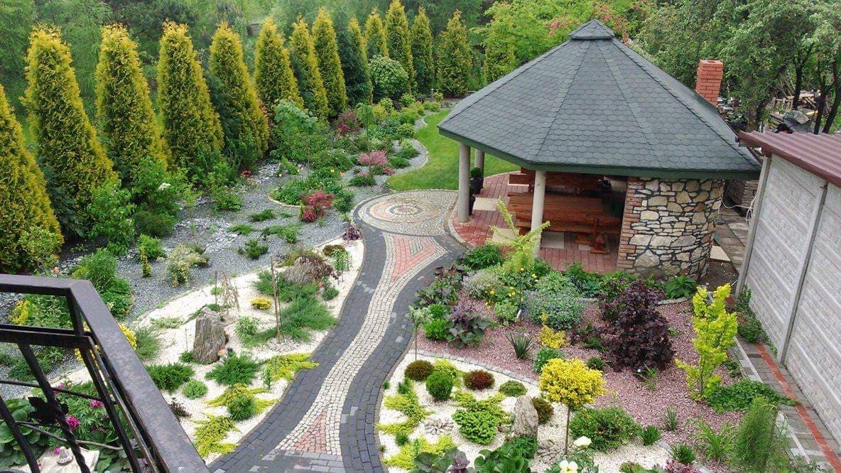 Widok z góry na starannie zaprojektowany ogród z wijącą się ścieżką z kostki brukowej w odcieniach szarości, czerwieni i bieli, prowadzącą do kamiennej altany z drewnianym wnętrzem, otoczonej...