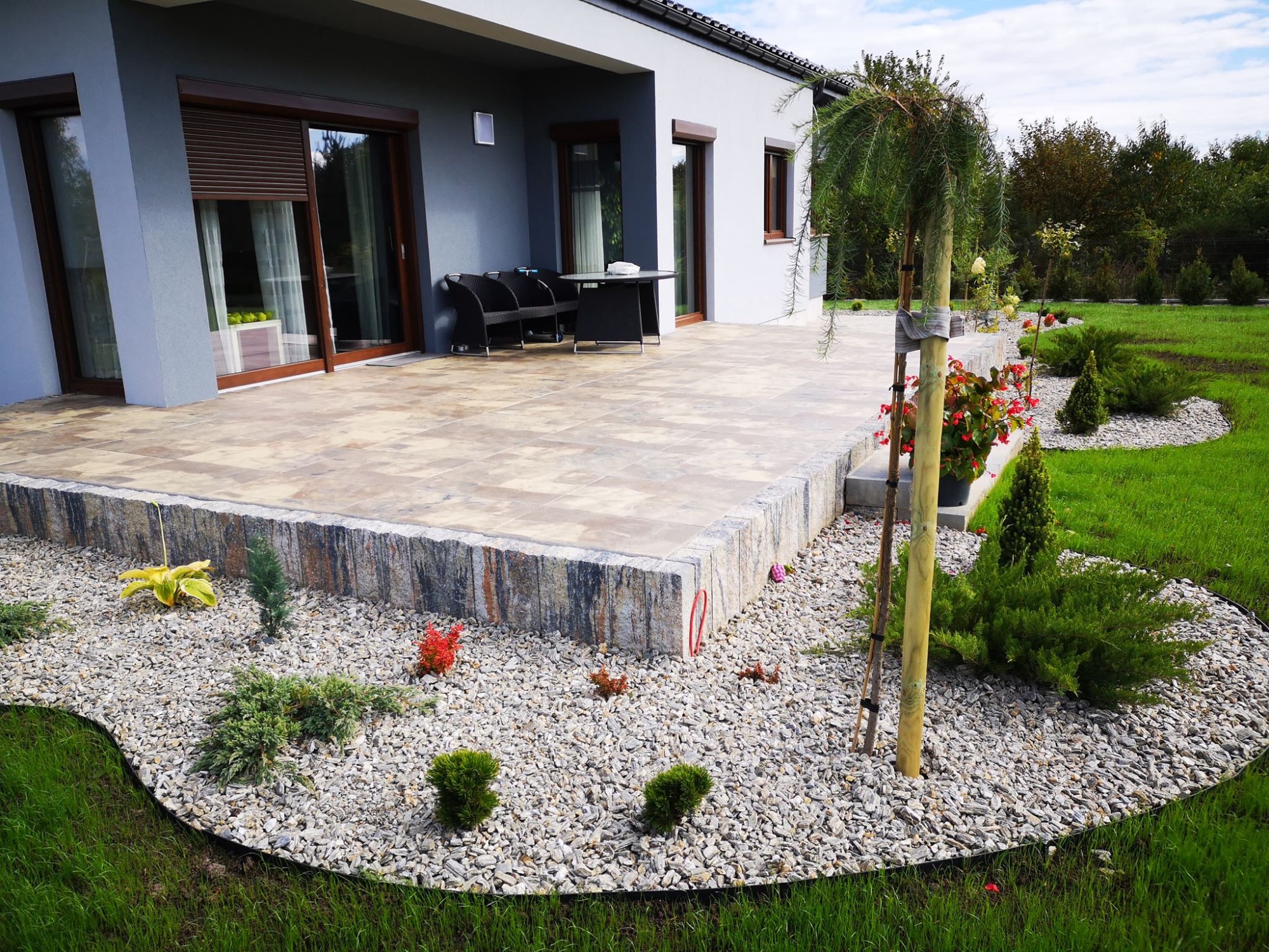 Nowoczesny taras z kamienną podmurówką, wyłożony jasnymi płytami, z widokiem na zadbany ogród z nasadzeniami i kamiennym podsypem.