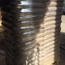 Stos worków z pelletem na palecie, owinięty folią stretch, z widocznym fragmentem otoczenia magazynowego.