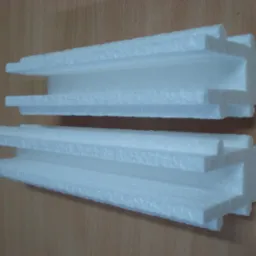 Dwa fragmenty białego styropianu o nieregularnych kształtach, ułożone jeden nad drugim na drewnianym tle, ukazujące strukturę materiału.