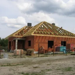 Budowa domu jednorodzinnego: surowa konstrukcja z czerwonej cegły z widoczną drewnianą więźbą dachową, palety z cegłami i materiałami budowlanymi na placu budowy, w tle fragment zabudowy sąsiedniej.