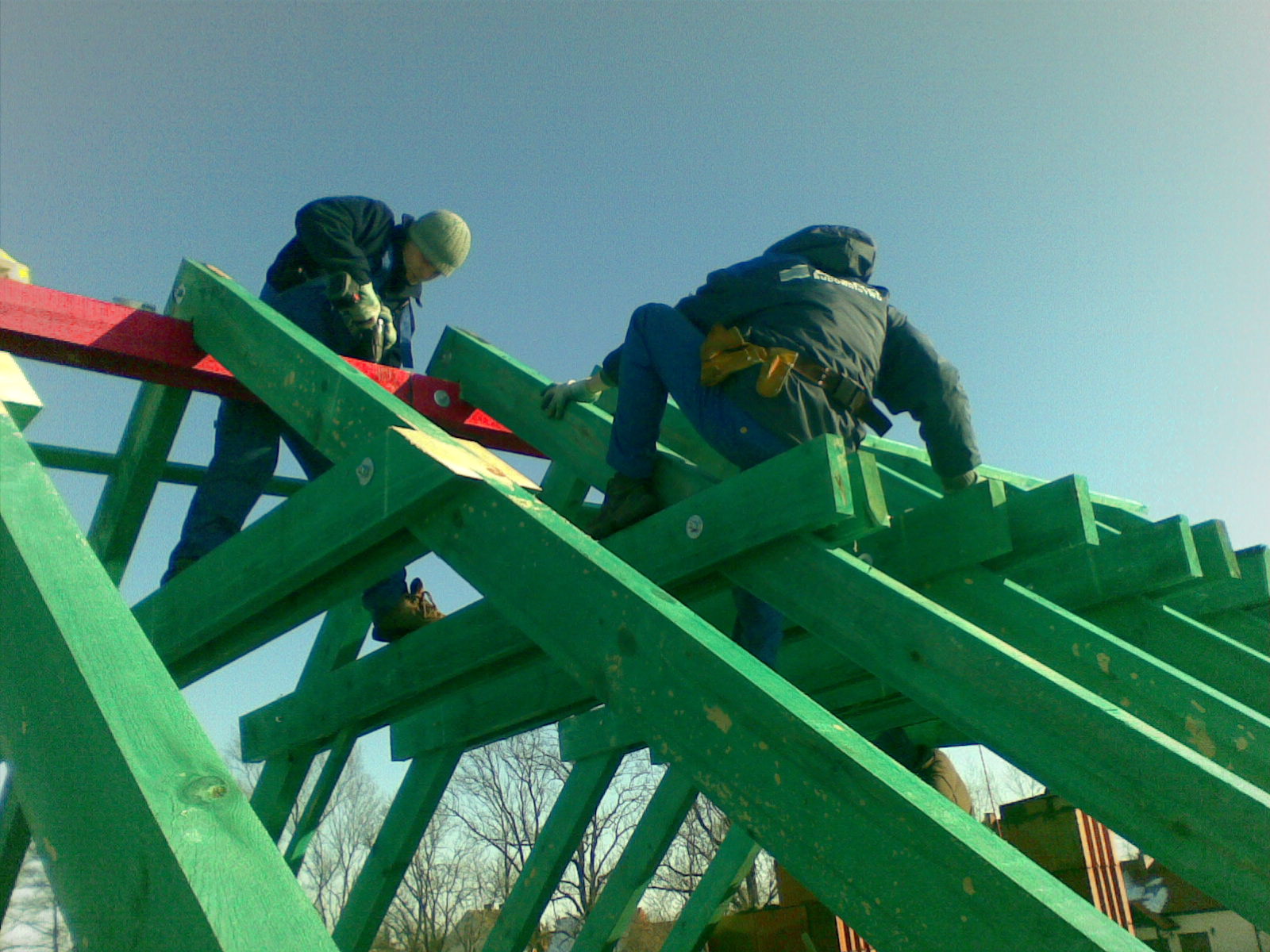 Dwóch dekarzy pracuje na zielonej więźbie dachowej z czerwonym elementem, widok z dołu na tle błękitnego nieba.