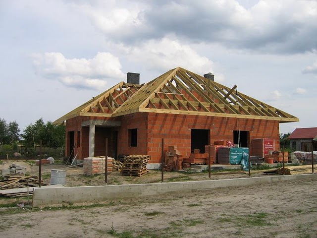 Budowa domu jednorodzinnego: surowa konstrukcja z czerwonej cegły z widoczną drewnianą więźbą dachową, palety z cegłami i materiałami budowlanymi na placu budowy, w tle fragment zabudowy sąsiedniej.