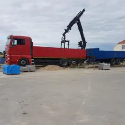 Czerwony samochód ciężarowy z dźwigiem i niebieską przyczepą, zaparkowany na placu budowy, obok palety z kostką brukową i worków z materiałami budowlanymi.