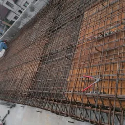 Siatka zbrojeniowa ułożona na drewnianym szalunku, widoczny pracownik w kombinezonie i kasku, używający narzędzi do mocowania pręt&oacute;w, w tle budynki mieszkalne.