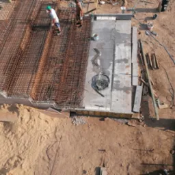 Widok z góry na plac budowy z rozległą siatką zbrojeniową, częściowo zabetonowaną płytą fundamentową, dwoma pracownikami w trakcie pracy i rozrzuconymi narzędziami na piaszczystym podłożu.