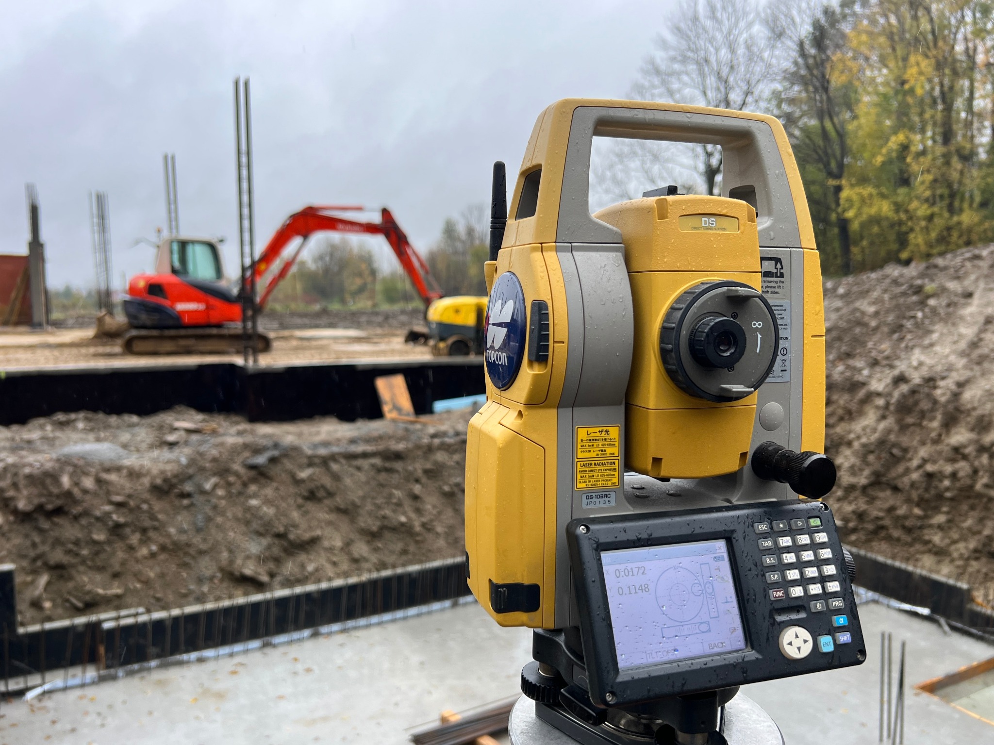 Żółty tachimetr Topcon DS na budowie w pochmurny dzień, w tle widoczne maszyny budowlane i zbrojenia fundamentów.