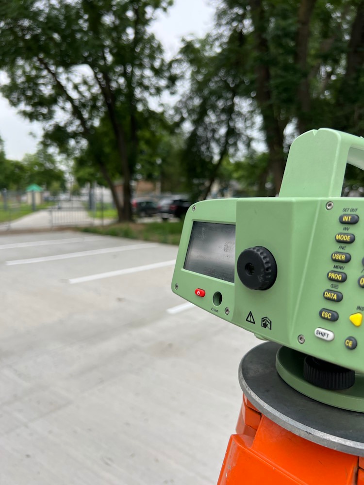 Zielony teodolit elektroniczny na pomarańczowym statywie ustawiony na betonowym parkingu z wyznaczonymi białymi liniami, w tle drzewa i zabudowania.